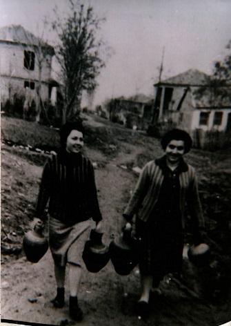 Δύο γυναίκες κουβαλούν στάμνες. Η μία φορά ζακέτα μαύρη μπεζ φούστα, κάλτσες και μαύρα παπούτσια. Η δεύτερη κοπέλα φοράει μαύρο φουστάνι, μαύρα παπούτσια και μπεζ ζακέτα. Ασπρόμαυρη φωτογραφία